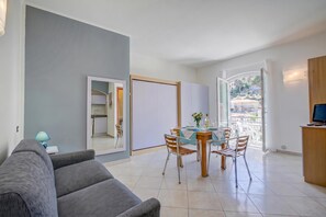 A modern living room with a grey sofa, a dining area with a glass table, and a view through a glass door.