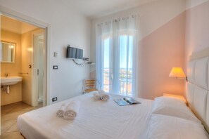 A hotel room with a bed, a TV, a window with curtains, a bedside lamp, and a bathroom with a sink and mirror. - Seaview Pool Retreat - M17, Andora, Italy (Andora)