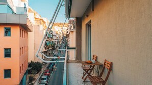 A narrow urban street with parked cars and a building with a balcony featuring two wooden chairs and a table. - Athens City Views - 4BR Luxury w/ Chef's Kitchen & Balconies (Athina)