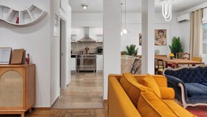 A modern living room with a yellow sofa, a blue armchair, and a wooden cabinet. A kitchen with stainless steel appliances is visible in the background. - Athens City Views - 4BR Luxury w/ Chef's Kitchen & Balconies (Athina)