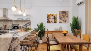 A modern kitchen with a marble island, wooden dining table, and chairs. There are potted plants and artwork on the walls. - Athens City Views - 4BR Luxury w/ Chef's Kitchen & Balconies (Athina)