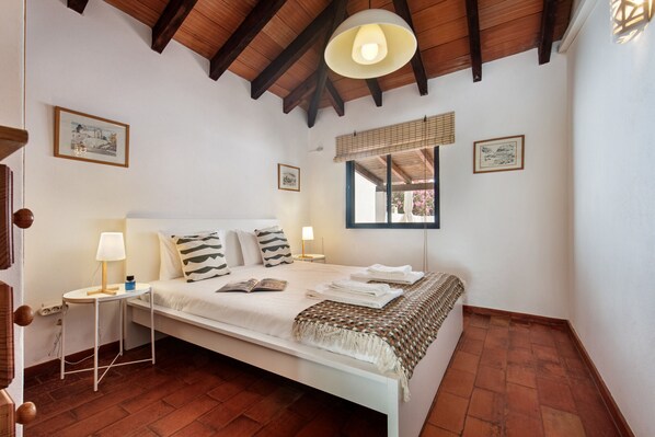 A bedroom with a white bed, a nightstand with a lamp, a window with blinds, and a ceiling fan.