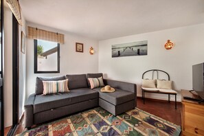 A modern living room with a grey sectional sofa, a patterned rug, a small wooden side table, a metal-framed bed, and a wall-mounted TV. - Retreat with Terrace and BBQ (Cabanas de tavira)