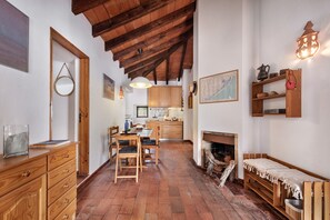 A rustic interior with a wooden dining table, chairs, a fireplace, and a kitchen in the background.