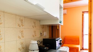 A kitchen with a stove, a kettle, and a microwave. There is a window with curtains and a bed with an orange blanket in the adjacent room. - Casa Anna by Interhome (Pompeiana)