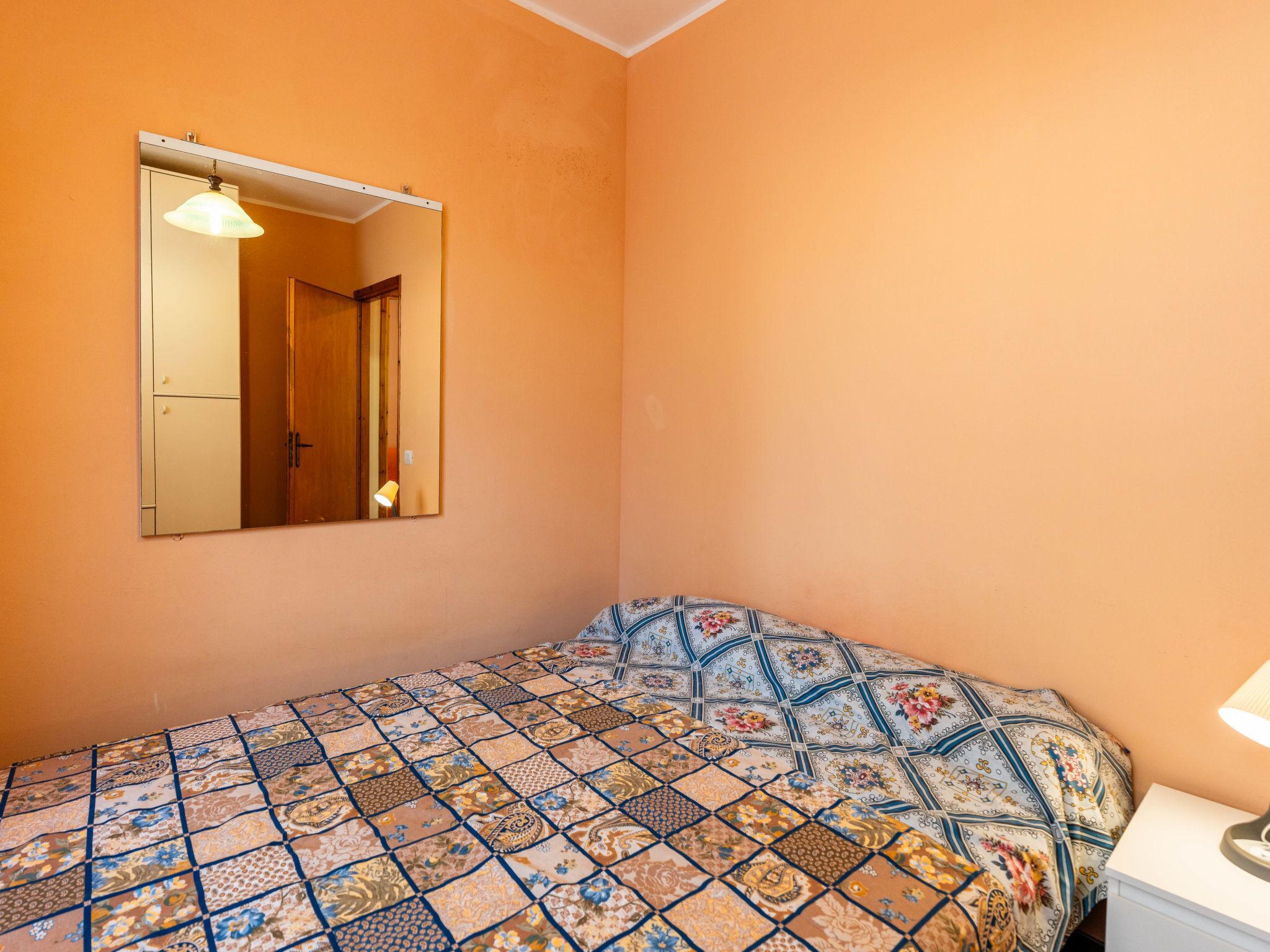 Ein Schlafzimmer mit einem gemusterten Bettbezug, einem Spiegel an der Wand und einem Nachttisch mit einer Lampe.