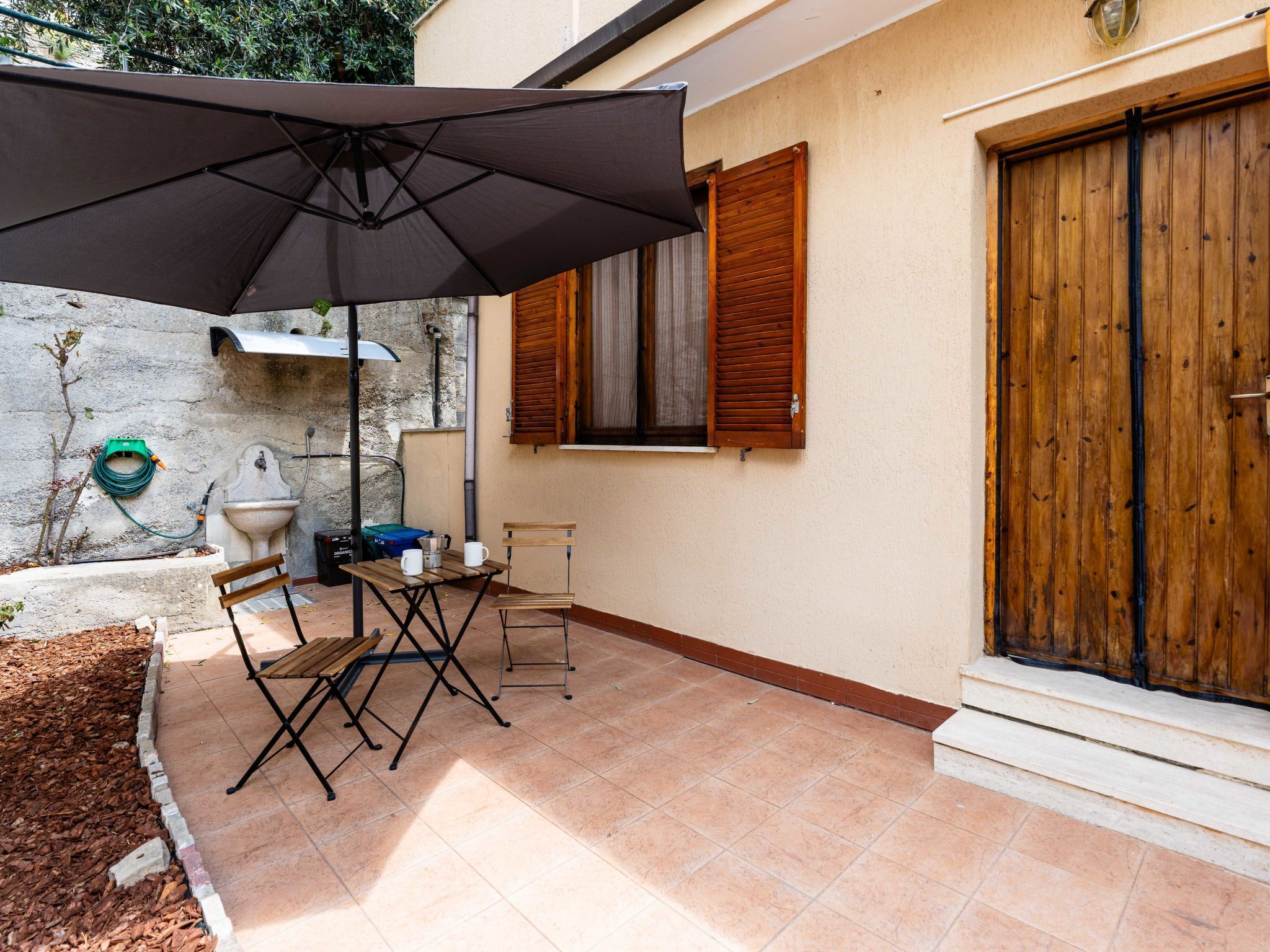 Eine Terrasse mit Tisch, Stühlen, einem großen Sonnenschirm, einer Springbrunnen und einer Holztür.