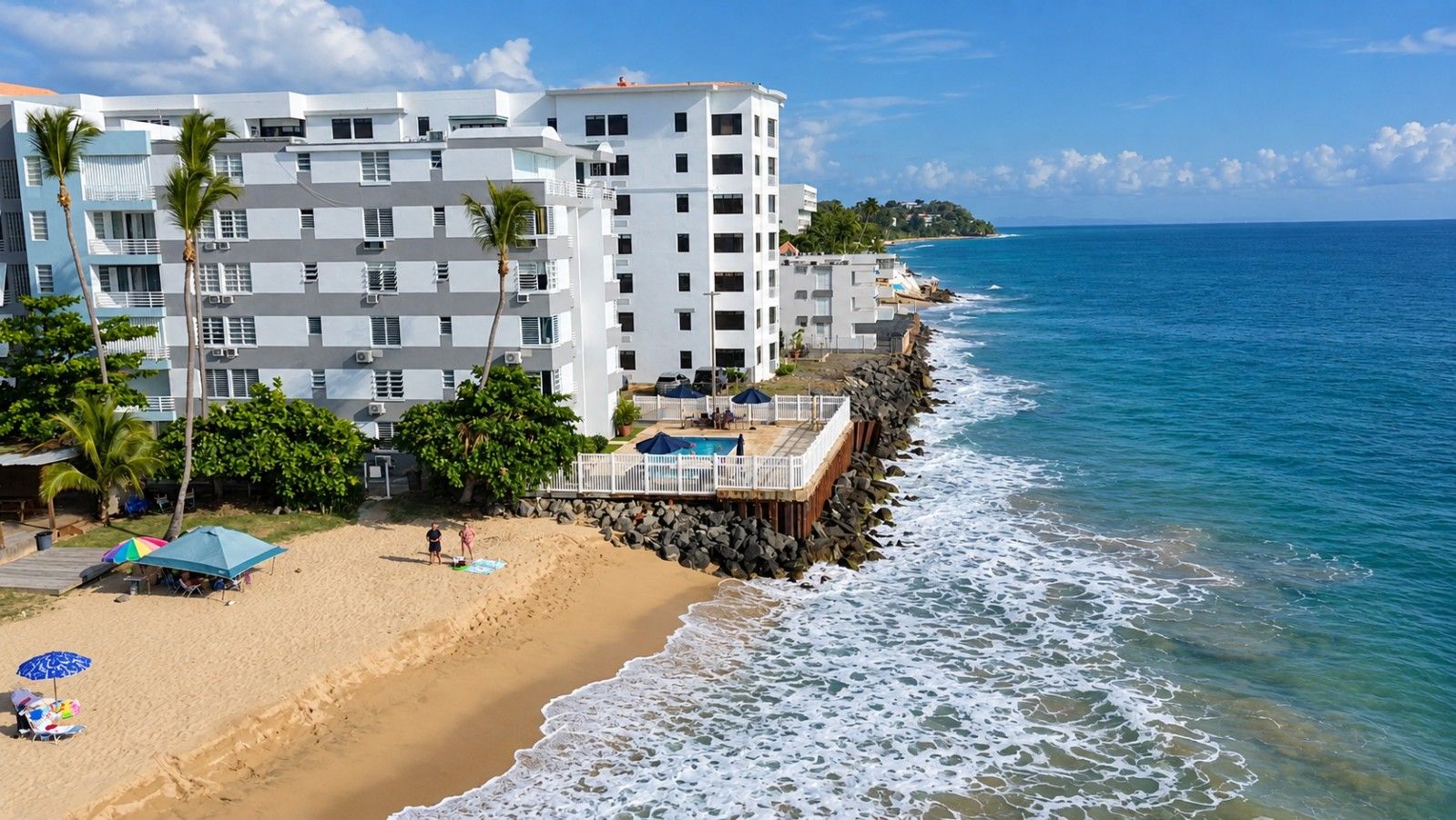 Un hotel frente a la playa con varios pisos, una playa arenosa y un rompeolas rocoso.