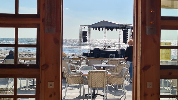 A wooden-framed window with a view of an outdoor seating area with tables and chairs, a stage with speakers, and a person standing.