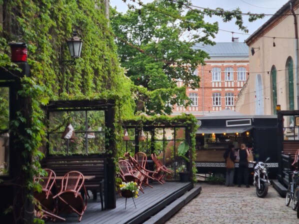 A cobblestone street with a covered outdoor seating area, wicker chairs, and a building with a brick facade in the background. - Cozy Studio in Liepāja by the sea (Liepaja)