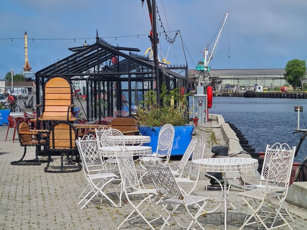 A waterfront café with outdoor seating, including metal chairs and tables, and a wooden structure providing shade.
