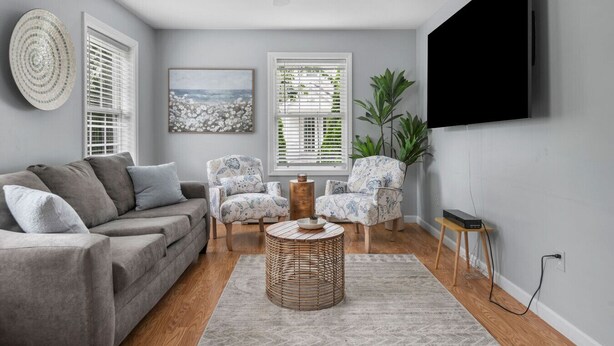 A living room with a grey sofa, two patterned armchairs, a round wicker coffee table, and a flat-screen TV mounted on the wall.