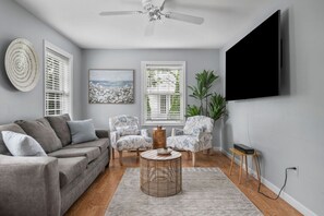 A living room with a grey sofa, two patterned armchairs, a round wicker coffee table, and a flat-screen TV mounted on the wall.