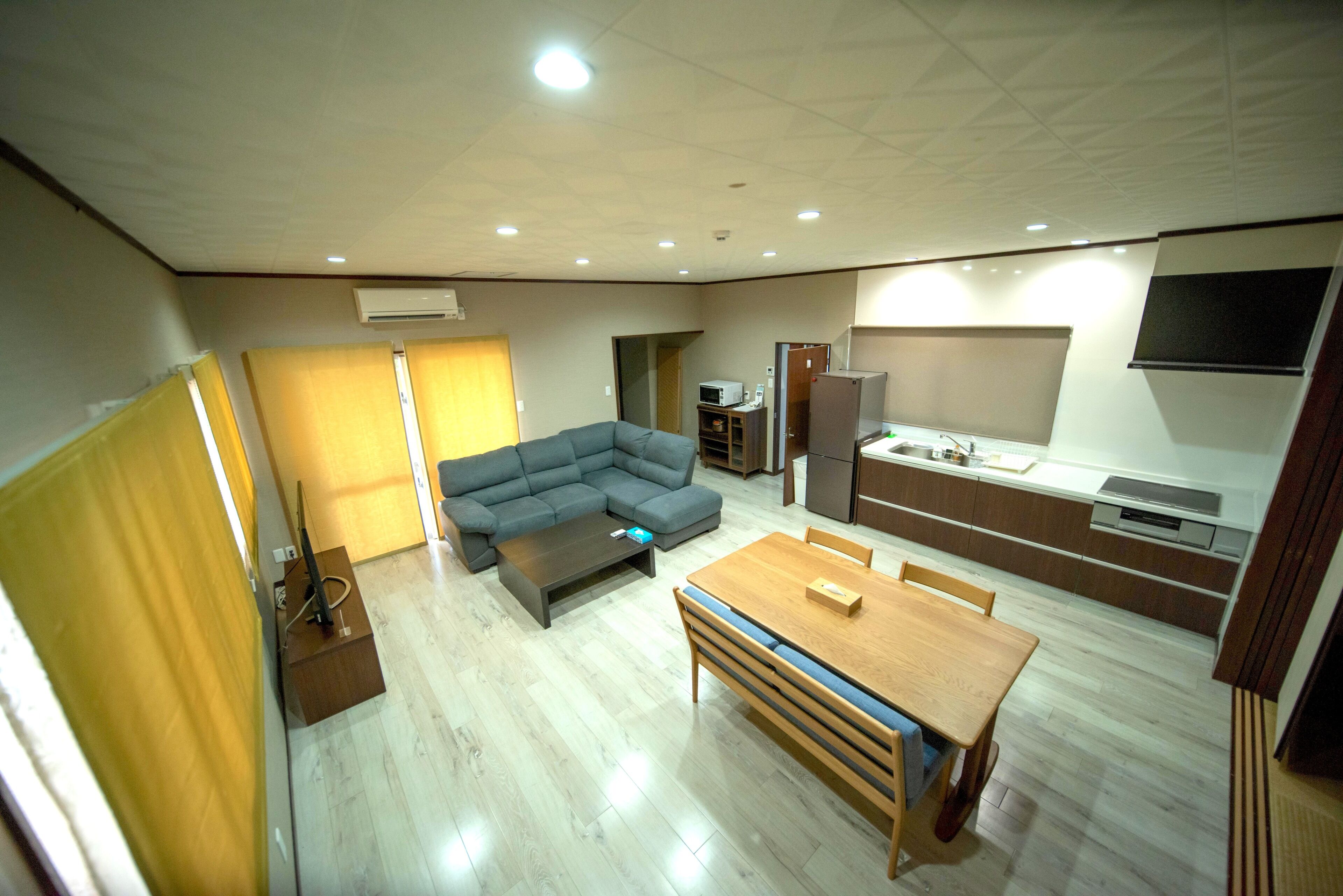 A modern living room with a sectional sofa, a wooden dining table, and a kitchen area with a microwave and refrigerator.