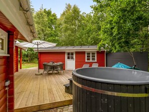 A wooden deck with a hot tub, a red building with a door and windows, and a table with chairs under an umbrella.