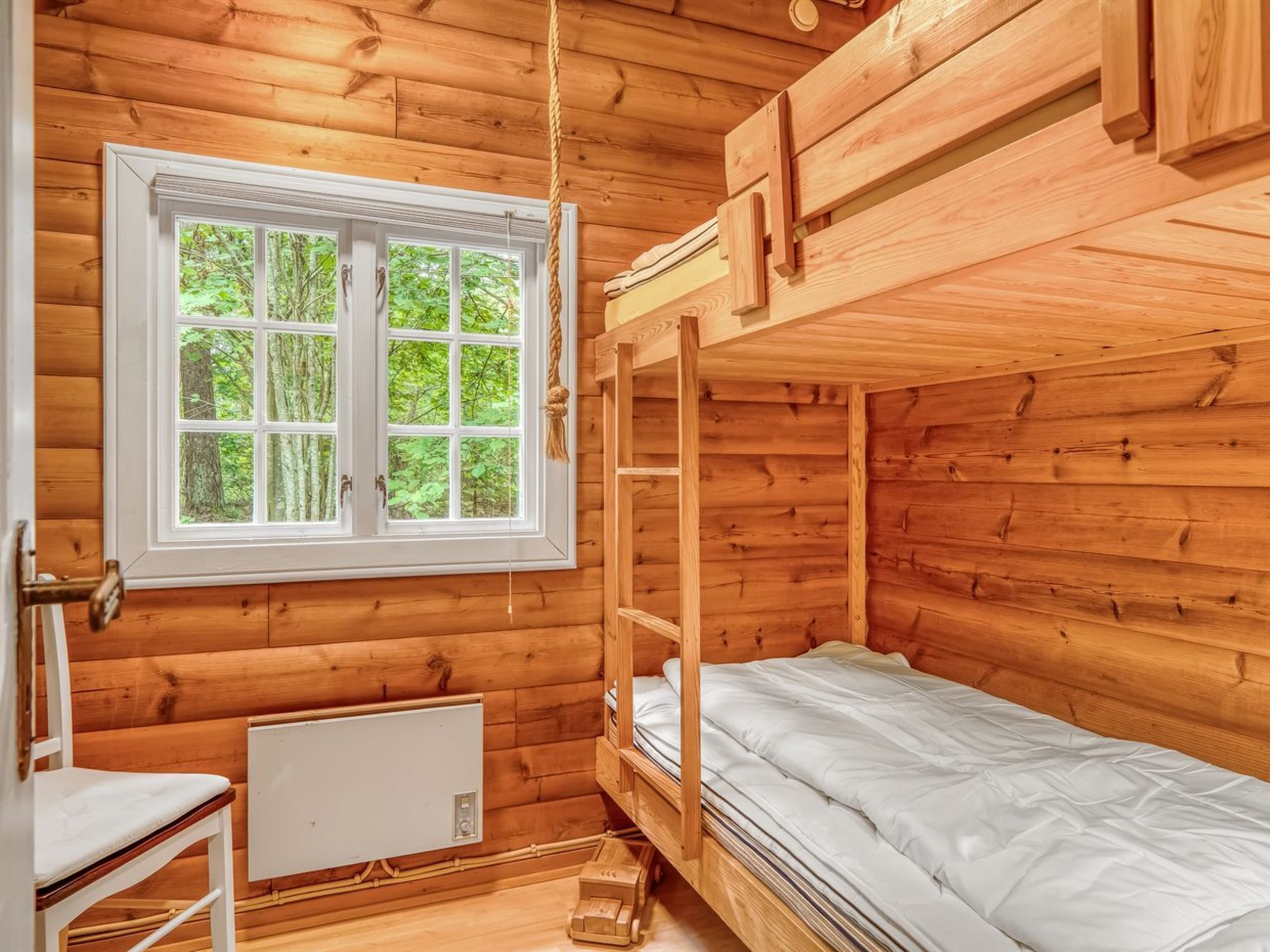 A wooden cabin room with a bunk bed, a window with a view of greenery, and a radiator.