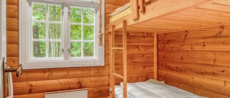 A wooden cabin room with a bunk bed, a window with a view of greenery, and a radiator.
