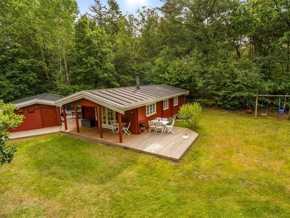 A wooden cabin with a covered patio, a deck, and a swing set in a wooded area.