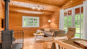L’intérieur chaleureux d’un chalet avec un poêle à bois, un canapé, une table à manger et de grandes fenêtres donnant sur un paysage verdoyant.