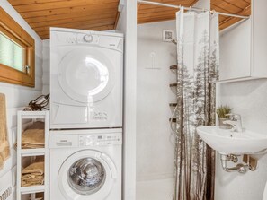 A compact laundry room with a top-loading washer and dryer, a shower with a forest-themed curtain, and a sink with a modern faucet.