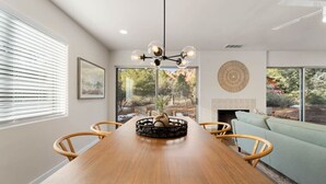 Un comedor con mesa de madera, sillas, una lámpara de araña y vistas al exterior a través de amplios ventanales.