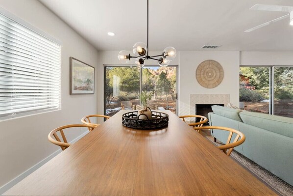 Un comedor con mesa de madera, sillas, una lámpara de araña y vistas al exterior a través de amplios ventanales.