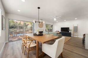 A spacious living room with a wooden dining table, comfortable seating, a large TV, and a view of the outdoors.