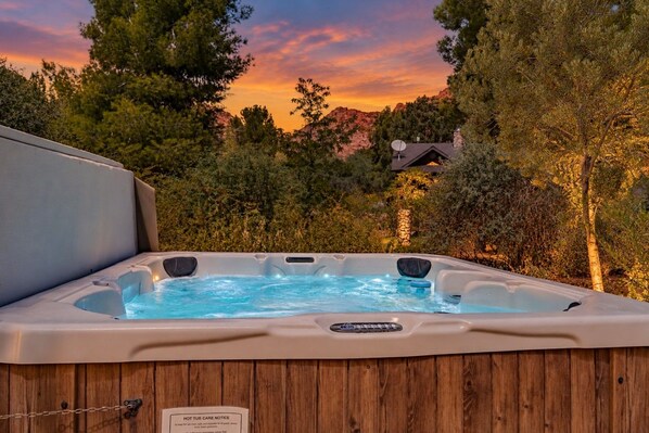 A hot tub in a wooden enclosure with a view of trees and a house in the background.