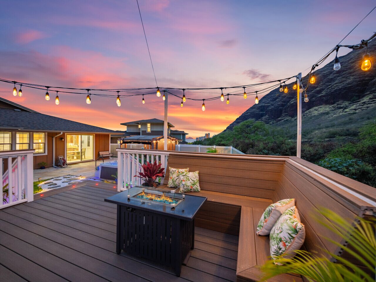 A deck with a fire feature, seating, and string lights against a sunset backdrop.