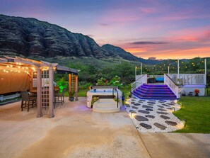 A hot tub with steps leading up to a deck, surrounded by a stone pathway and a covered seating area with string lights. - Single LVL Home|Beach 5 Min|Fire Pit|Free Parking (Waianae)