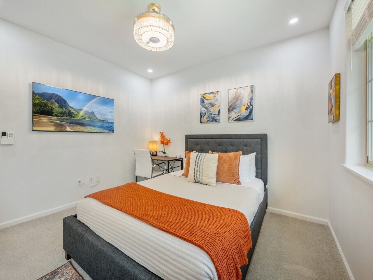 A modern bedroom with a large bed, a desk, and a chair. There are two paintings on the wall and a chandelier above the bed.