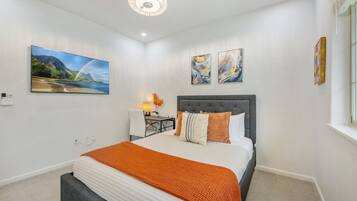 A modern bedroom with a large bed, a desk, and a chair. There are two paintings on the wall and a chandelier above the bed.