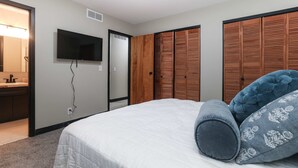Une chambre à coucher comprenant un lit, un téléviseur fixé au mur, une armoire en bois et une salle de bain avec un miroir et un lavabo.