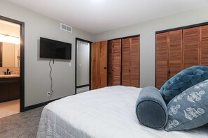 Une chambre à coucher comprenant un lit, un téléviseur fixé au mur, une armoire en bois et une salle de bain avec un miroir et un lavabo.