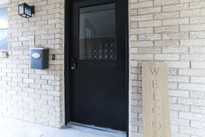 Une porte noire avec une pancarte indiquant « Bienvenue », une boîte aux lettres et un luminaire fixé au mur.