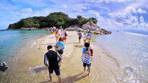 A beach with clear water, people walking, and a rocky island in the background.