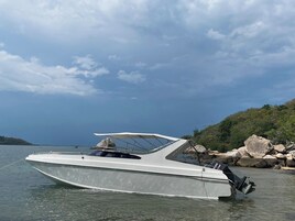 Un barco de motor blanco con cabina, amarrado cerca de una costa rocosa bajo un cielo despejado.