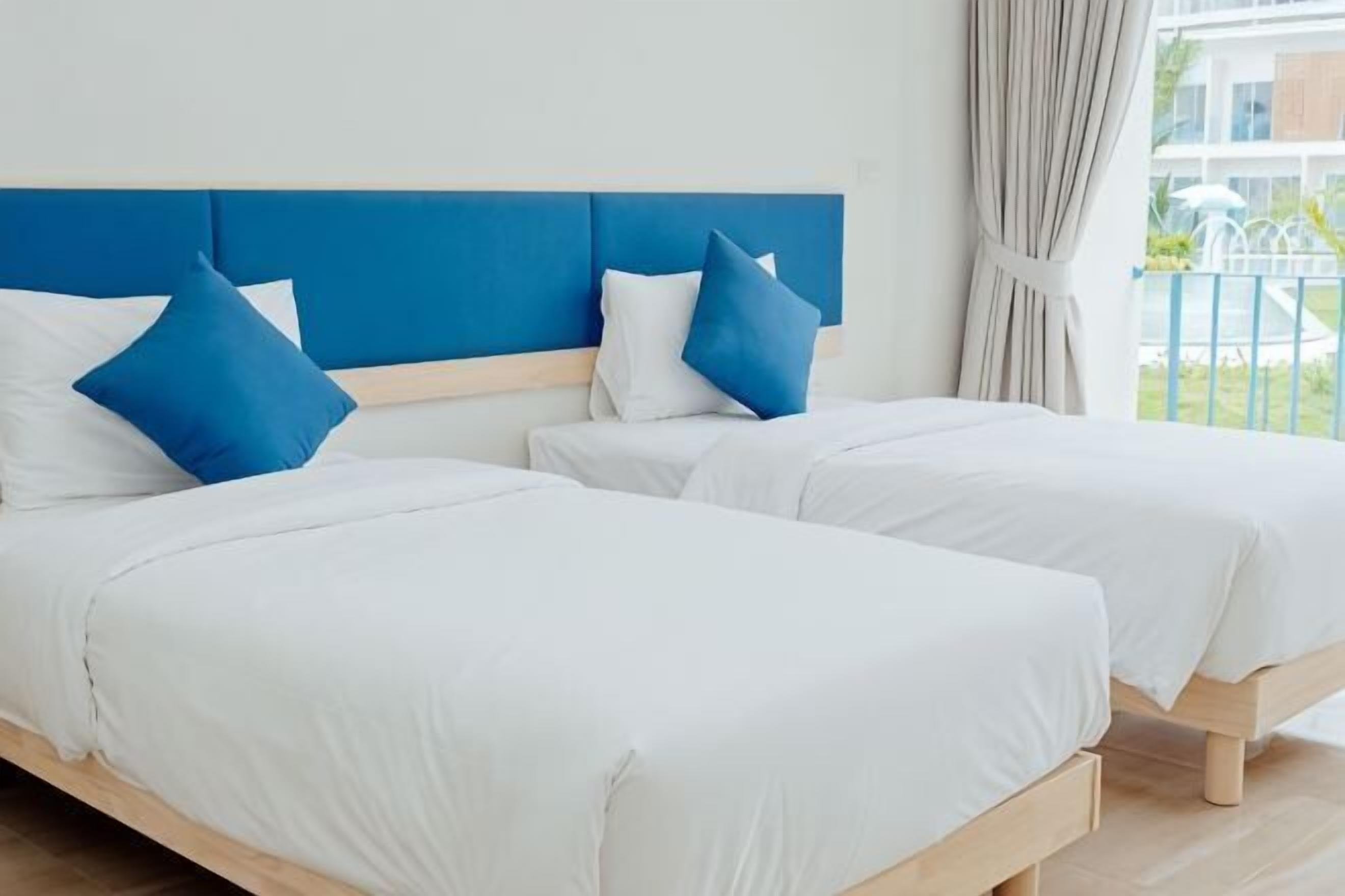 Dos camas individuales con ropa blanca y almohadas azules, un cabecero azul y vistas a un balcón con barandilla.
