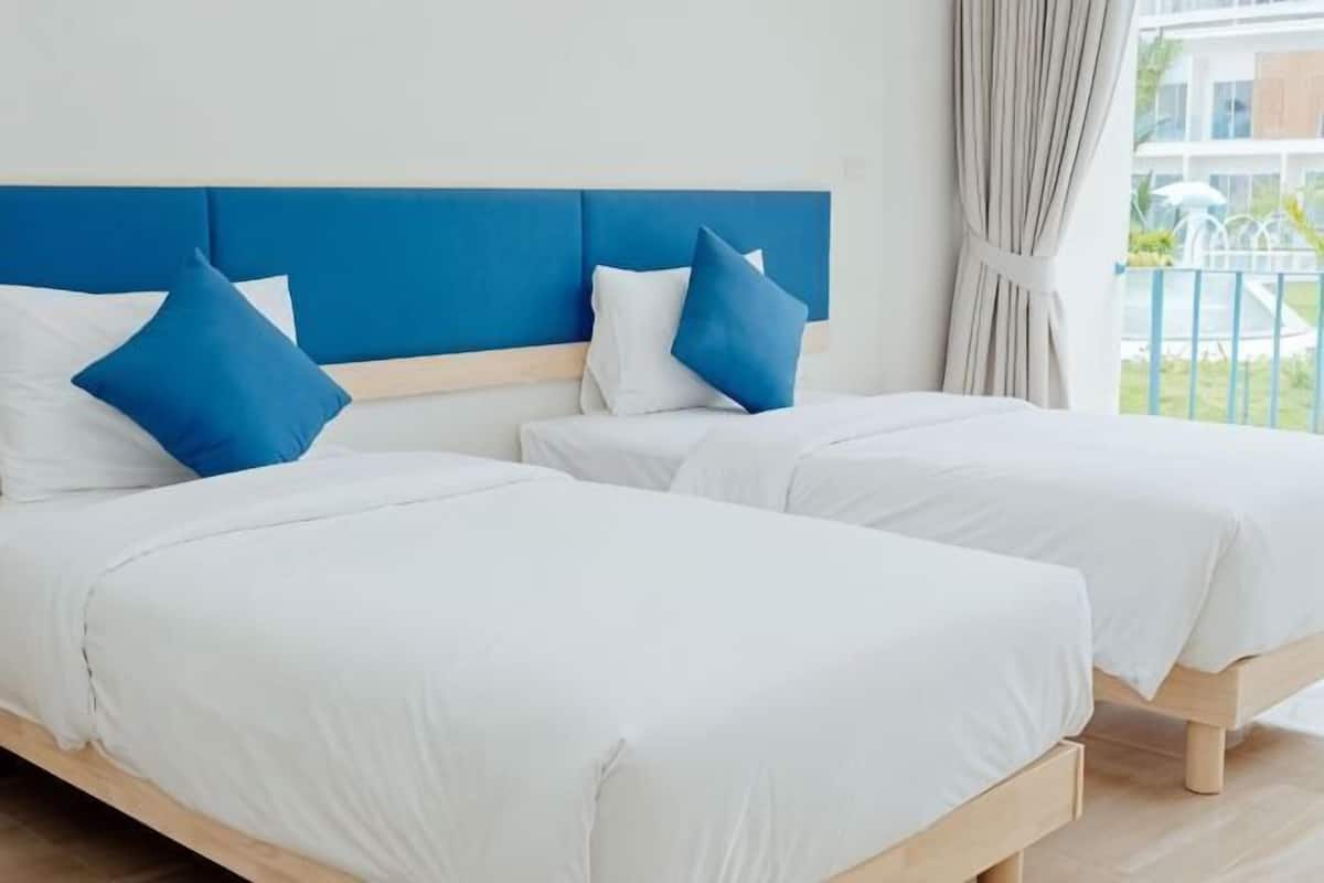 Dos camas individuales con ropa blanca y almohadas azules, un cabecero azul y vistas a un balcón con barandilla.