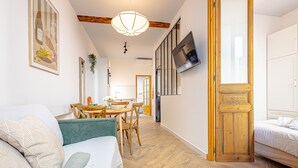 Una habitación moderna y bien iluminada, con piso de madera, un comedor con mesa y sillas, y una cama junto a una puerta de madera.