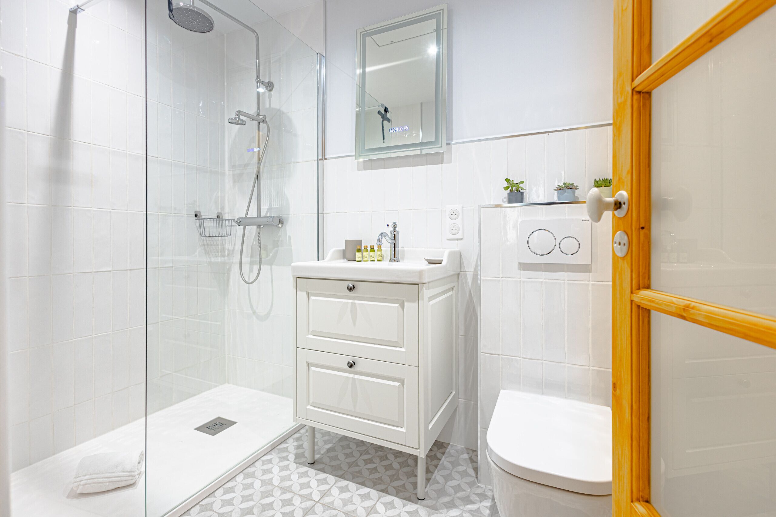 A modern bathroom with a glass shower enclosure, a white sink with a mirror above, and a wooden door.