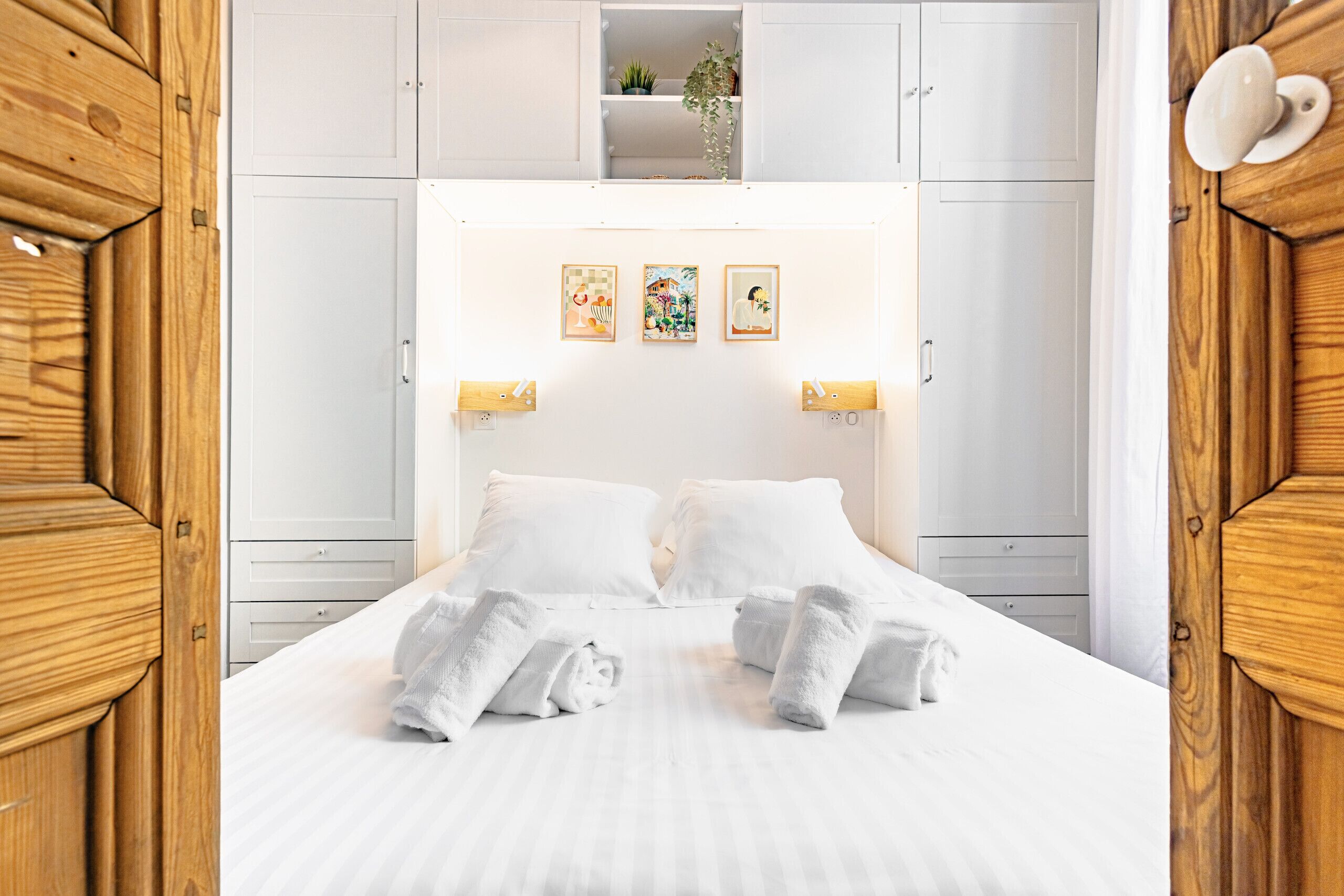 A neatly made bed with white linens and towels, a white headboard with wall-mounted lights, and a shelf above the bed with a plant.