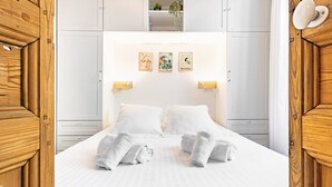 Una cama bien hecha con ropa de cama y toallas blancas, un cabecero blanco con luces fijadas a la pared y una estantería sobre la cama con una planta.