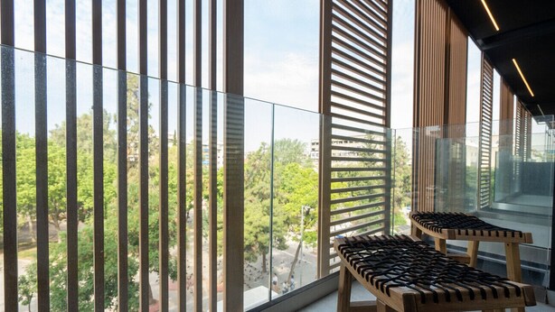 A balcony with a view of trees and buildings, wooden benches, and vertical blinds.