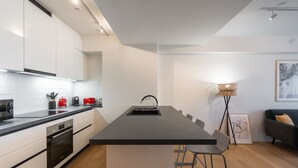 A modern kitchen with white cabinets, a black countertop, and a central island with a sink. There are two bar stools, a floor lamp, and a framed picture on the wall.
