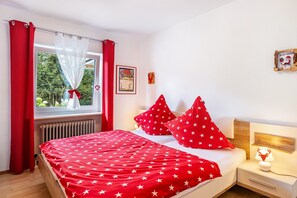 Un dormitorio con cama, cortinas rojas, ventana con vistas, lámpara de noche y un adorno en la pared.