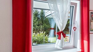 Una ventana con cortina blanca atada con un lazo rojo, un radiador abajo y vista a zonas verdes al exterior.