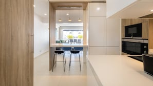 A modern kitchen with a white countertop, two bar stools, a built-in oven, and pendant lighting.