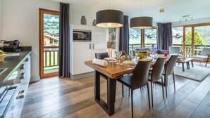 A modern dining area with a wooden table, black chairs, and pendant lights. A kitchen with a microwave and cabinets is visible in the background. - APARTMENT  LA  CHAPELLE D'ABONDANCE - Oasis B107 (LA CHAPELLE D ABONDANCE)