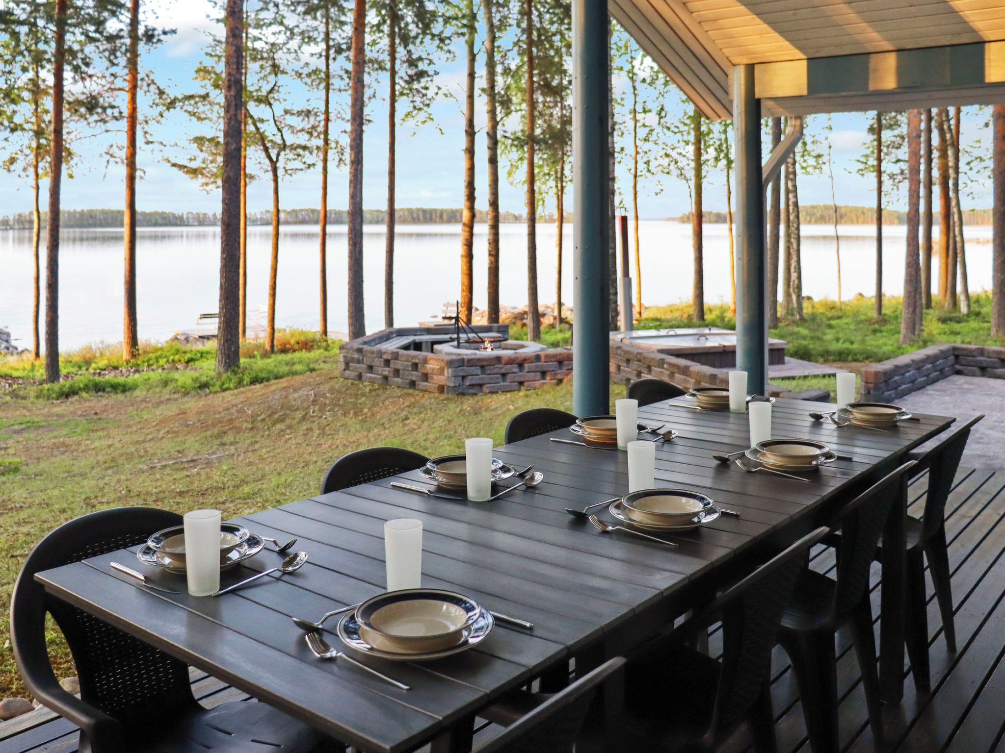 Uma área de refeições ao ar livre com uma mesa comprida posta para seis pessoas, rodeada por cadeiras, com vista para um lago, árvores e um poço de fogueira de pedra ao fundo.